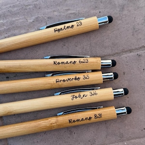 May include: Five bamboo pens with silver accents and black stylus tips. Each pen is engraved with a different Bible verse: "Psalms 23", "Romans 6:23", "Proverbs 3:5", "John 3:16", and "Romans 8:31". The pens are light brown.