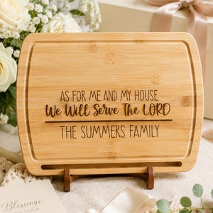 May include: A rectangular bamboo cutting board with rounded corners and a juice groove. The board is engraved with the text AS FOR ME AND MY HOUSE We Will Serve The LORD and THE SUMMERS FAMILY. It rests on a small wooden stand for display.