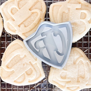 May include: A gray plastic cookie cutter in the shape of a shield with a stylized letter "T" inside. The cutter is on top of a cooling rack with several sugar cookies that have been cut out with the same cutter.