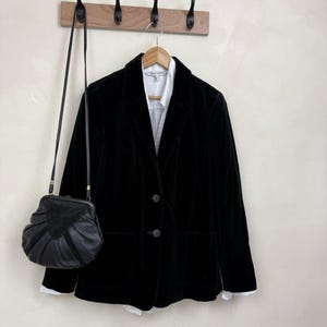 Peut inclure: Un blazer en velours noir avec deux boutons sur le devant et deux poches, superposé sur une chemise blanche à col. Un sac à main en cuir noir avec une fermeture à cadre métallique et une longue bandoulière est suspendu à côté du blazer.