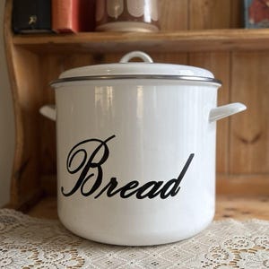 May include: A white cylindrical enamel bread bin with a matching lid and two side handles. The word Bread is printed on the front in a black cursive script. The container features a thin silver-coloured metal rim around the top edge.