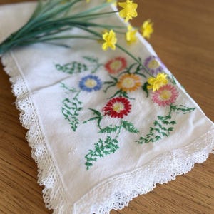 Peut inclure: Mouchoir blanc brodé avec un motif floral coloré et des feuilles vertes, bordé d'une délicate dentelle blanche. Un bouquet de jonquilles jaunes repose sur le mouchoir, créant un thème printanier.