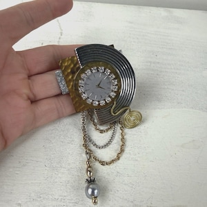 May include: A vintage brooch with a watch face, silver dial, and crystal detailing. The brooch has a mixed-metal design with gold, silver, and bronze tones, and dangling chains with a grey pearl. The brooch is held in a hand.