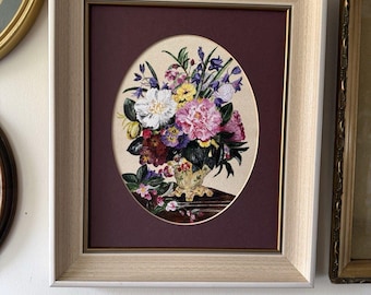 Op de afbeelding: Een ingelijst borduurwerk met een kleurrijk boeket bloemen in een decoratieve vaas. Het ovale handwerk toont witte, roze, gele en paarse bloemen met groene bladeren, geplaatst tegen een dieprode passe-partout in een lichthouten lijst.