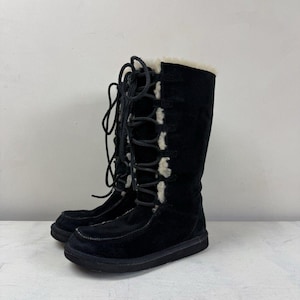 May include: A pair of black mid-calf boots featuring a soft cream-coloured faux fur lining that extends along the front lace-up closure. The boots have black laces, a rounded toe box, and a flat black sole, providing a sturdy and functional design.