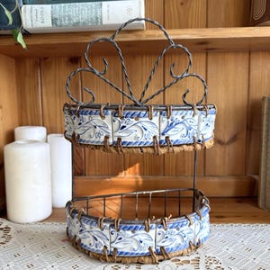 Peut inclure: Étagère décorative à deux niveaux avec une structure en fer forgé. Les étagères sont bordées de carreaux à motifs floraux bleus et blancs et de garnitures en paille tressée. L'étagère est conçue pour contenir de petits objets.