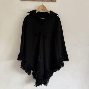 Pode incluir: Um poncho preto com capuz apresentando uma bainha pontiaguda com franjas longas nas bordas. A peça inclui um capuz com cordões pretos correspondentes e detalhes sutis de bordado tonal na superfície frontal do tecido.
