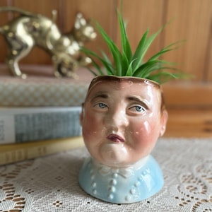 Könnte beinhalten: Ein Vintage-Keramik-Pflanzgefäß in Form eines menschlichen Kopfes mit blauem Kragen und Fliege. Der Pflanztopf hat ein bemaltes Gesicht mit rosigen Wangen und blauen Augen und enthält eine grüne Pflanze. Eine goldene Katzenstatue befindet sich im Hintergrund.