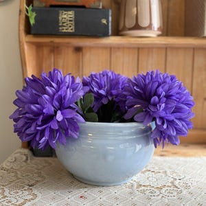 May include: A light blue ceramic bowl filled with three large, vibrant purple chrysanthemum flowers. The bowl has a smooth, glossy finish and a rounded shape, providing a stark colour contrast against the deep purple petals of the artificial blooms.