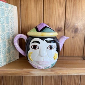 Può includere: Una teiera in ceramica con un design a volto. La teiera presenta un volto nero con grandi occhi, una luna crescente e una stella. Il coperchio è viola, verde e giallo. Il manico e il beccuccio sono viola chiaro. Su una mensola di legno.