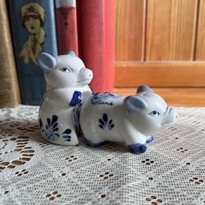 May include: Two white ceramic pig ornaments with blue detailing, including ears, snouts, and hooves, are displayed on a lace cloth. One pig has a blue bow tie. The word "Holland" is written on one pig.