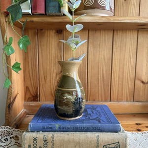 May include: A small, handcrafted ceramic vase with a landscape design, holding a sprig of faux eucalyptus. The vase is placed on top of two vintage books, with a wooden shelf and other books in the background. The vase is approximately 5 inches tall.