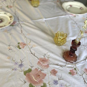 May include: A white tablecloth with embroidered pink roses, green leaves, and delicate purple flowers. The table is set with vintage plates, yellow candle holders, and a decorative candle holder with a red base.