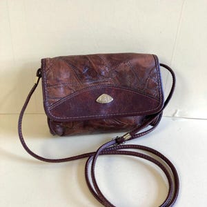 May include: A vintage, brown leather crossbody bag with a flap closure. The bag features a stitched design and a long, thin strap. A small, gold-colored emblem is on the front. The bag is a deep burgundy color.