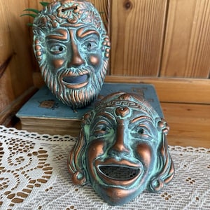May include: Two antique-style theatrical masks with a weathered copper and green patina. One mask depicts a bearded man, the other a smiling woman. The masks are likely made of a cast metal and are displayed on a wooden surface.