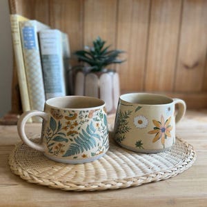 May include: Two ceramic mugs with a light beige base color. The left mug features a dense pattern of green ferns and small orange and yellow wildflowers. The right mug displays a spaced-out design of white daisies and yellow coneflowers.