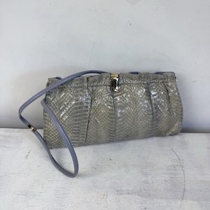 May include: A light grey, textured clutch bag with a matching shoulder strap. The bag has a rectangular shape with a gold-coloured clasp and a gathered design. The strap is a light blue-grey colour.