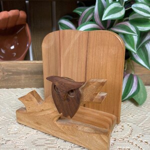 Op de afbeelding: Een houten servethouder met een gesneden uil op een tak. De uil is gemaakt van donkerder hout, wat contrasteert met het lichtere hout van de houder en de tak. De houder heeft een rechthoekige achterkant en een gleuf voor servetten.