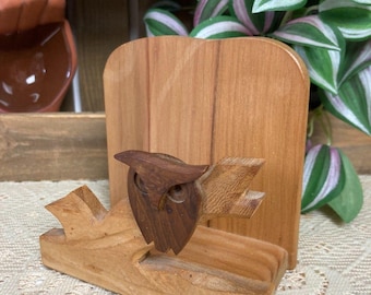 May include: A wooden napkin holder with a carved owl perched on a branch. The owl is made of a darker wood, contrasting with the lighter wood of the holder and branch. The holder has a rectangular back and a slot for napkins.