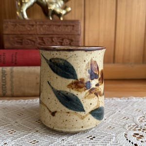 May include: A ceramic mug with a beige speckled background and a brown rim. The mug is decorated with dark blue and brown leaf and floral patterns. The mug is sitting on a white lace doily.