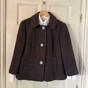 May include: A dark brown jacket featuring a repeating pattern of small white dots, a wide collar, and three large light blue buttons down the front. The jacket is layered over a white collared shirt with long sleeves that extend past the cuffs.