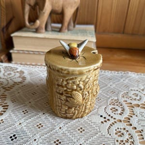 Könnte beinhalten: Ein zylindrischer Honigtopf aus Keramik mit glänzender, hellbrauner Glasur und einer strukturierten Wabenoberfläche. Der Deckel ist mit einer modellierten Bienenfigur mit bemalten Flügeln und braunem Körper versehen und hat eine Aussparung.