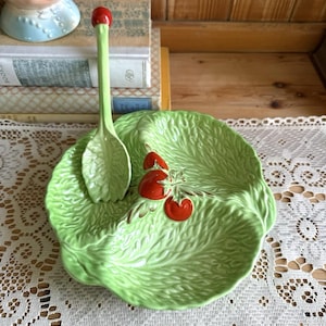 May include: A light green ceramic serving dish shaped like a lettuce leaf with three red tomato accents in the centre. The dish includes a matching light green spoon with a red handle tip, featuring a textured leaf design on the bowl of the spoon.