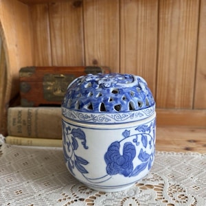 Könnte beinhalten: Ein weißes Keramikgefäß mit blauen Blumenmustern und einem abnehmbaren Deckel. Der Deckel hat ein durchbrochenes Design mit kleinen Blumenmotiven. Der zylindrische Körper ist mit handgemalten blauen Ranken und Blüten verziert.