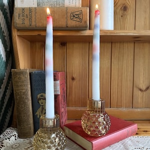 Op de afbeelding: Twee brandende taps toelopende kaarsen met kleurrijke strepen in gouden glazen houders. De houders staan op een rood boek. Een verzameling vintage boeken en een witte pilaarkaars staan op de achtergrond.