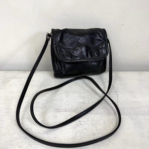 May include: Black leather crossbody bag with a flap closure. The bag has a textured surface and a long, thin strap. The bag is designed to be worn over the shoulder or across the body. The bag is a small size, suitable for carrying essentials.