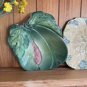 Op de afbeelding: Een groene keramische blad-vormige serveerschaal met een getextureerd oppervlak en een roze centrum. De schaal wordt tentoongesteld op een houten plank met een ander bord en gele bloemen.