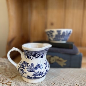 May include: A white ceramic creamer pitcher featuring a blue willow pattern with detailed illustrations of pagodas, bridges, and birds. The pitcher has a curved handle and a pouring spout, showcasing a classic transferware design in blue and white.