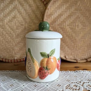 Puede incluir: Un tarro de cerámica blanco con tapa y pomo verde. El tarro está decorado con ilustraciones coloridas de frutas, incluyendo una pera, un melocotón y una fresa. El tarro está sobre un mantel de encaje blanco.