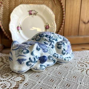 Può includere: Statuina di gatto in ceramica in posizione di riposo, con motivi floreali e a foglie blu su base bianca. Il gatto è su una tovaglia di pizzo bianco, con un piatto decorativo e un cestino di vimini sullo sfondo.