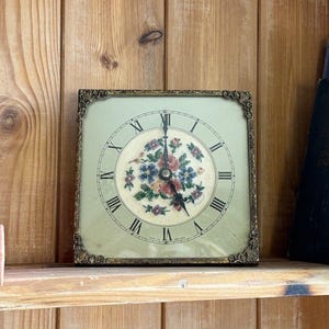May include: A square wall clock with a light green face and a central circular floral embroidery featuring red, blue, and green thread. The clock has black Roman numerals, thin black hands, and an ornate, dark metallic frame with corner details.