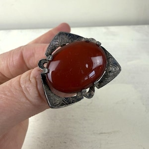 Op de afbeelding: Een vintage zilveren ring met een grote, ovale, gepolijste, roodbruine steen. De steen is gezet in een decoratieve zilveren zetting met bladvormige details. De ring is een statement piece en toont een gedurfd ontwerp.