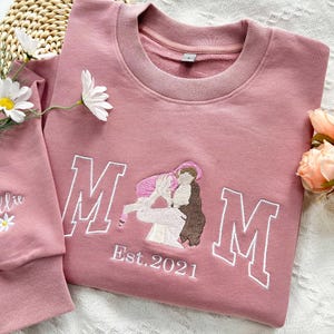 May include: Pink sweatshirt with embroidered white letters spelling "MOM" and a small embroidered image of a mother holding a child. The text "Est. 2021" is embroidered below the letters. The sweatshirt has a white embroidered word "Ollie" on the left sleeve.
