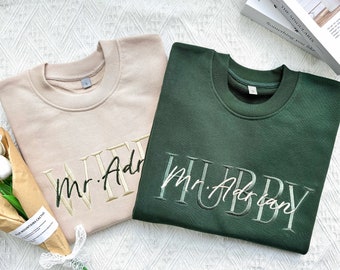 May include: Two sweatshirts, one beige and one dark green, with the words "Wife" and "Hubby" embroidered on them. The text also includes "Mr. Adrian" on both sweatshirts.