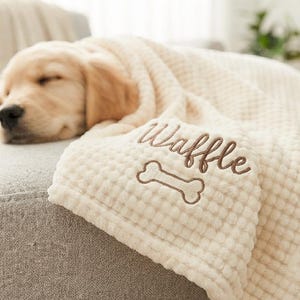 May include: A cream-coloured waffle-textured blanket featuring the embroidered name Waffle in brown script lettering above a brown embroidered dog bone icon. The blanket is draped over a sleeping golden-coloured puppy on a grey surface.