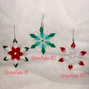 May include: Three glass snowflake ornaments in red, white, and green. Snowflake #1 is red and white with green accents. Snowflake #2 is green and teal. Snowflake #3 is red and white. Each has a silver wire hanger.