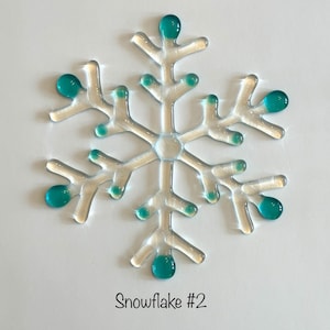 May include: A clear glass snowflake ornament with teal blue accents. The snowflake is a six-pointed star shape with intricate details. The ornament is labeled "Snowflake #2".