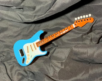 May include: A blue and white electric guitar with a brown neck and white knobs. The guitar is lying on a black fabric surface.
