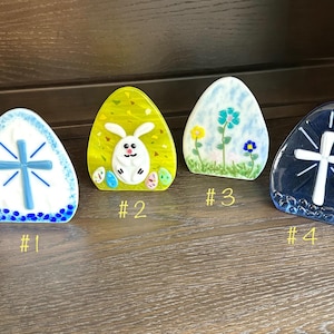 May include: Four glass Easter egg-shaped ornaments with different designs. The first ornament is white with a blue cross, the second is yellow with a white bunny, the third is blue with flowers, and the fourth is blue with a white cross.