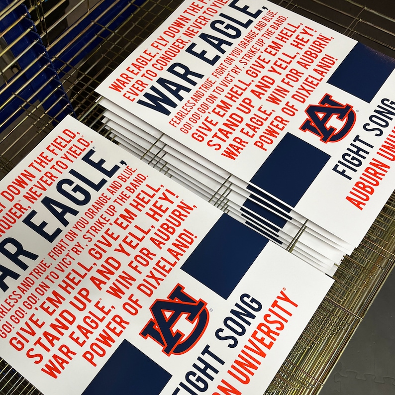 K&ouml;nnte beinhalten: Ein Stapel wei&szlig;er Schilder mit blauem und orangefarbenem Text. Die Schilder zeigen das Logo der Auburn University und die Worte "War Eagle", "Give 'em Hell", "Stand Up and Yell, Hey!" und "Fight Song".