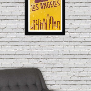 May include: A framed poster with a black border, featuring a basketball and the words "LOS ANGELES" in a distressed font. Below the text is a stylized skyline in a matching color, set against a yellow background. The poster is hanging on a white brick wall.