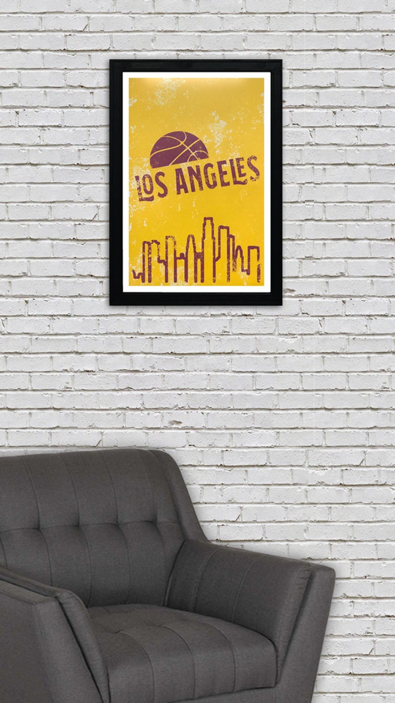May include: A framed poster with a black border, featuring a basketball and the words "LOS ANGELES" in a distressed font. Below the text is a stylized skyline in a matching color, set against a yellow background. The poster is hanging on a white brick wall.