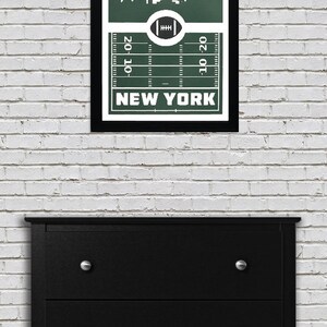 Puede incluir: Una impresi&oacute;n enmarcada con un fondo verde que presenta una silueta blanca del horizonte de la ciudad de Nueva York. El horizonte est&aacute; sobre un campo de f&uacute;tbol con las palabras "NEW YORK" impresas debajo del campo.