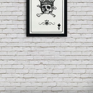 May include: A framed black and white illustration of the Ace of Spades playing card. The card features a skull wearing a crown and crossed bones. The card is framed in a black frame and is hanging on a white brick wall.