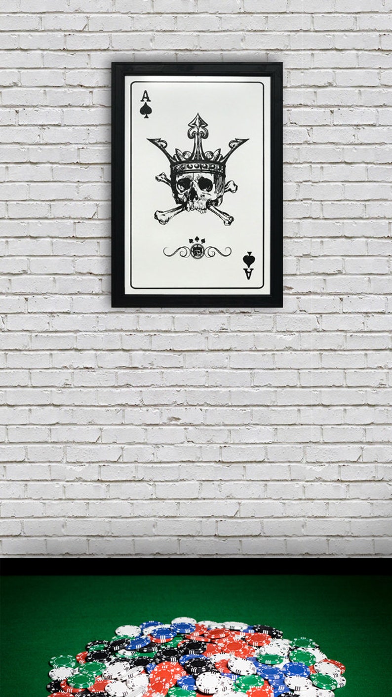 May include: A framed black and white illustration of the Ace of Spades playing card. The card features a skull wearing a crown and crossed bones. The card is framed in a black frame and is hanging on a white brick wall.