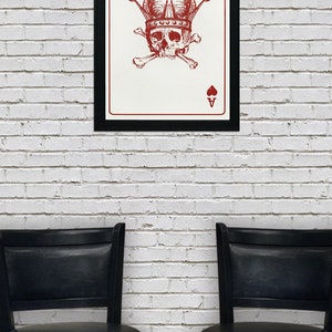 May include: A framed art print featuring a red skull wearing a crown, crossed bones, and the Ace of Spades and Hearts symbols. The artwork is displayed on a white brick wall, with two black chairs below.